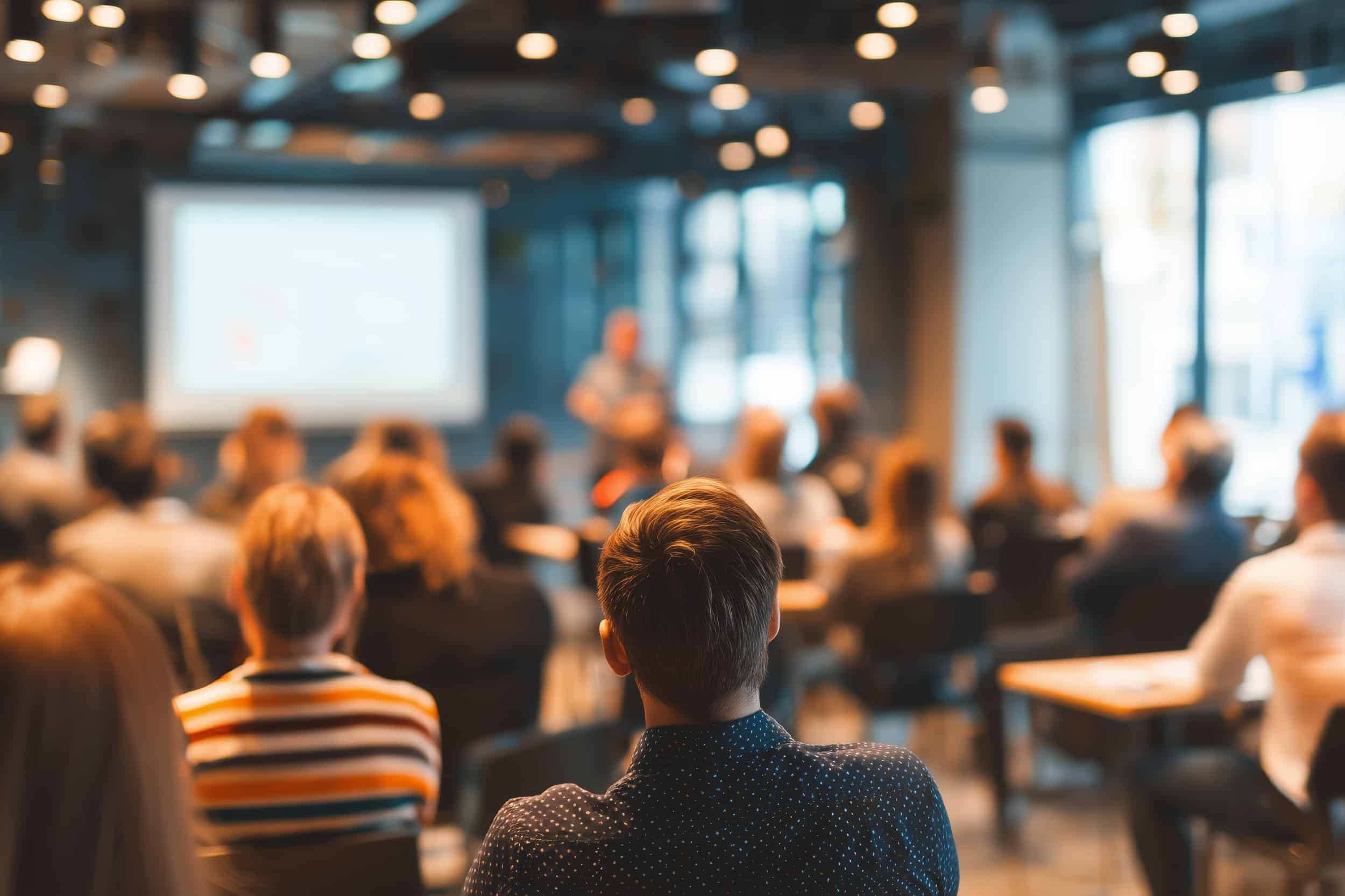 Audience seated in a modern conference room watching a presenter at the front with a blurred projector screen in the background.