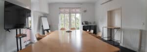 Bright conference room with a long wooden table and beige chairs, glass doors at the far end, and a wall-mounted TV on the left.