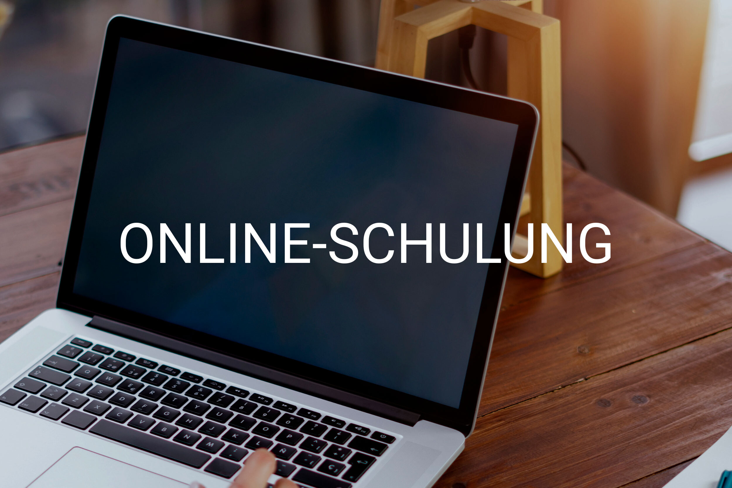 Laptop on a wooden desk displaying the text 'ONLINE-SCHULUNG' on the screen, suggesting online training.