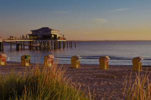 Beach at sunset with a wooden pier and pavilion over calm water; striped beach chairs line the sandy foreground.